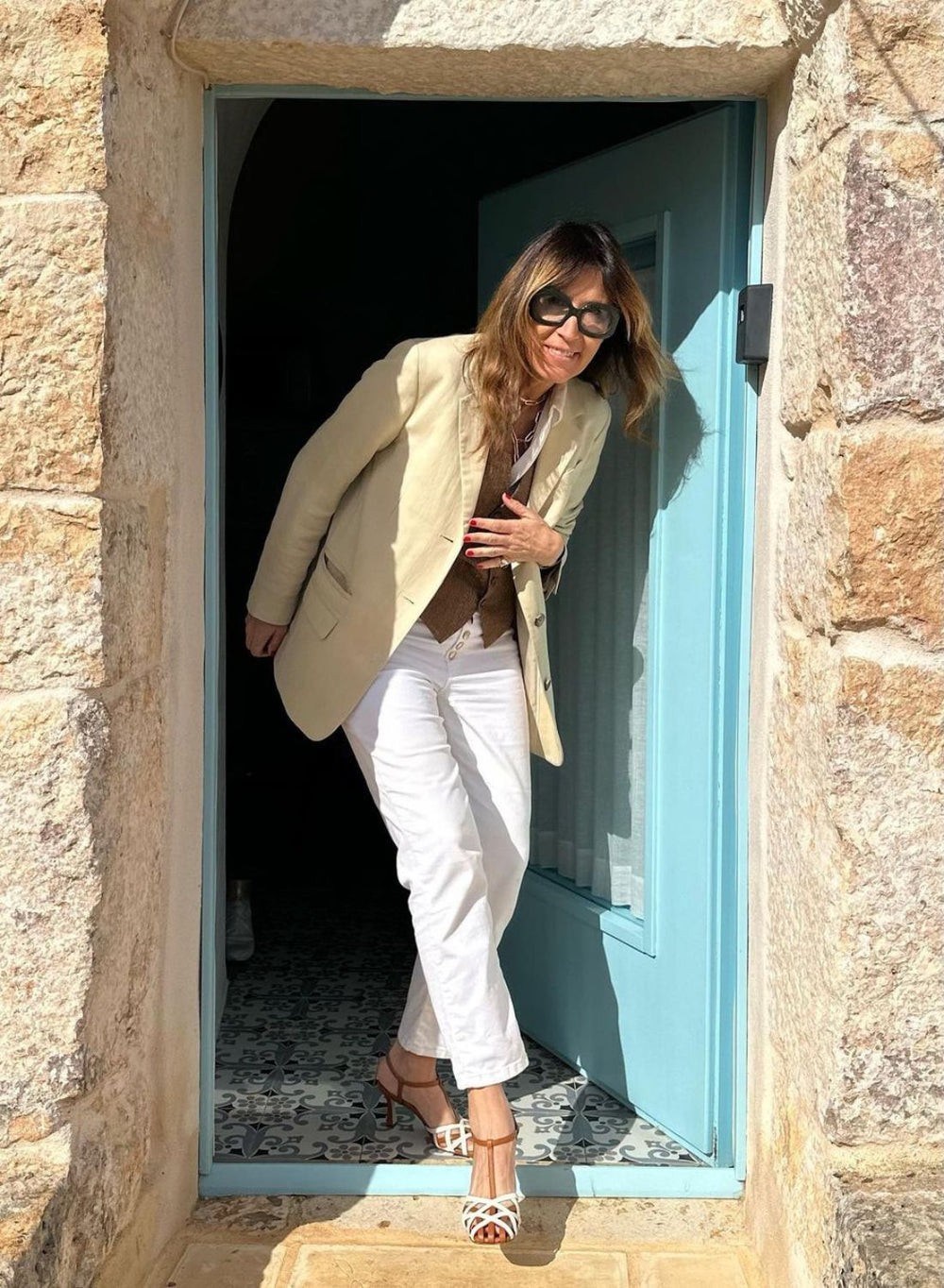 Simona sorride sulla soglia di una porta azzurra, in blazer chiaro e jeans bianchi